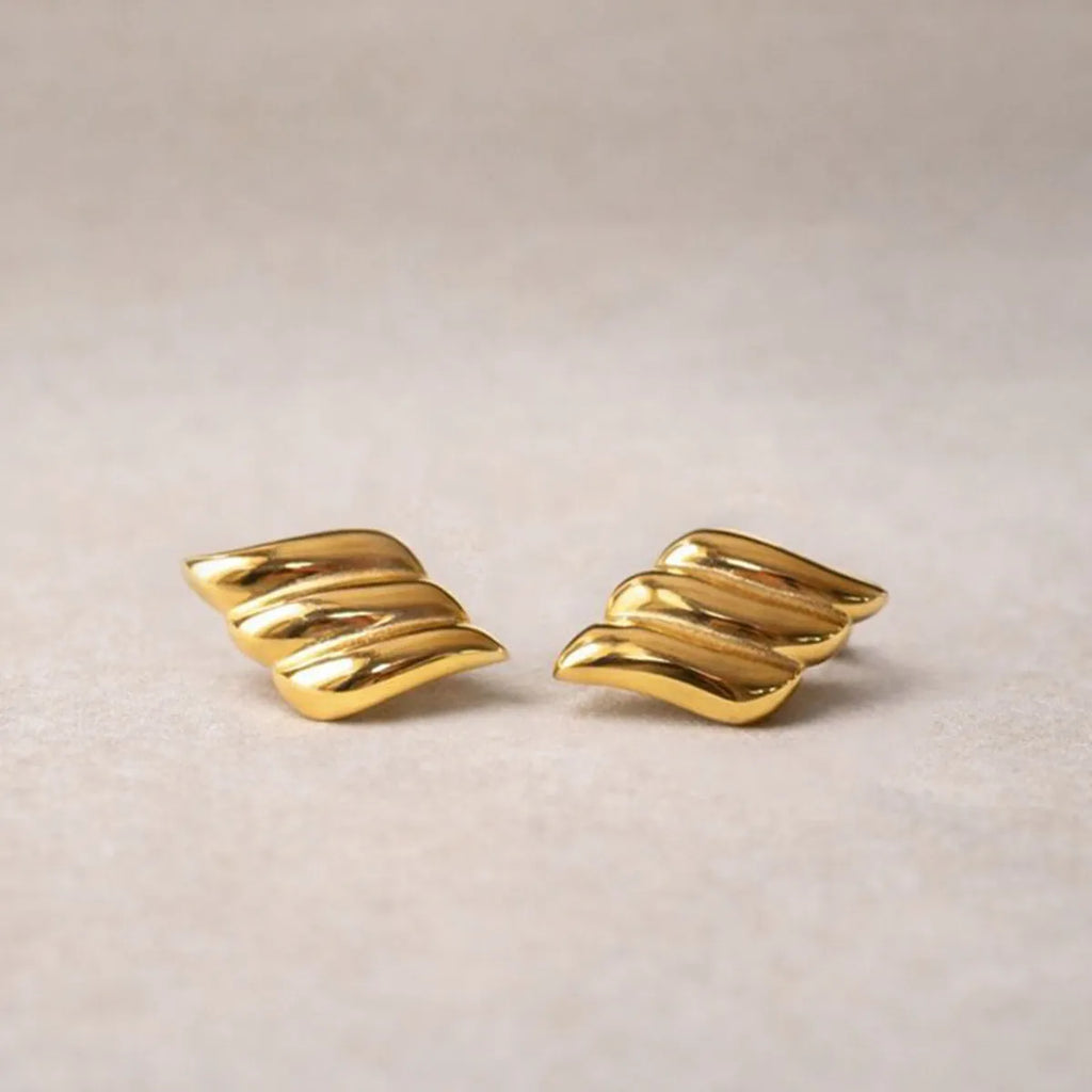 Boucles d'oreilles Envolée, ailes dorées en acier inoxydable,  sur un fond texturé beige clair.