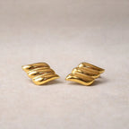 Boucles d'oreilles Envolée, ailes dorées en acier inoxydable,  sur un fond texturé beige clair.