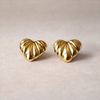 Boucles d'oreilles Passionnée en acier inoxydable doré, au design en relief en forme de cœur, posées côte à côte sur un fond beige texturé.