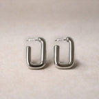 Boucles d'oreilles Franche, créoles rectangulaires en acier inoxydable argenté, photographiée en vue rapprochée,  sur une toile beige texturée.