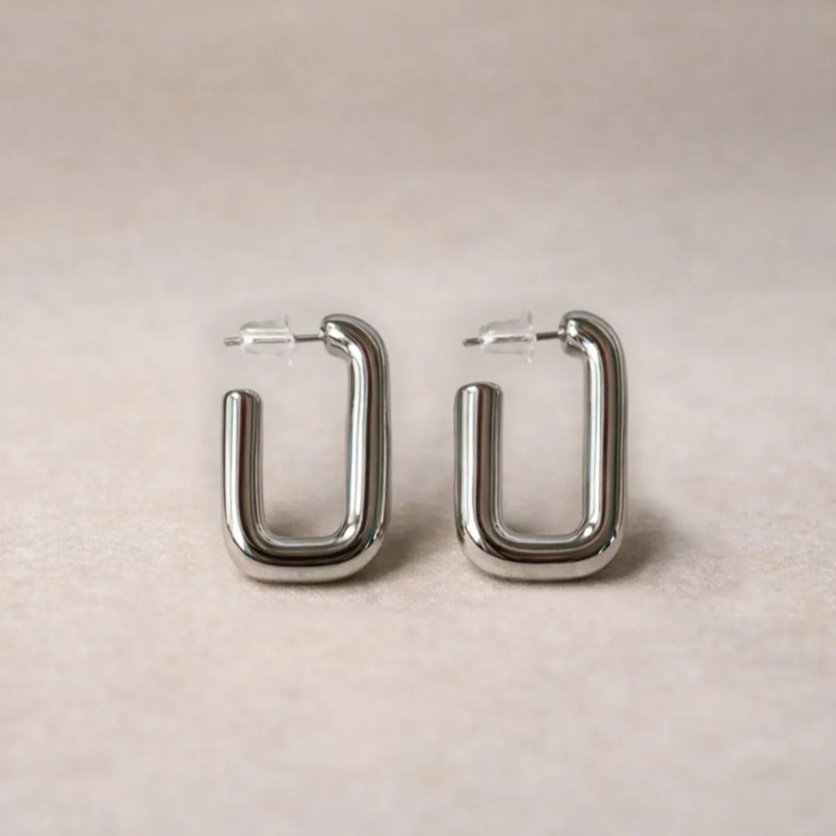 Boucles d'oreilles Franche, créoles rectangulaires en acier inoxydable argenté, photographiée en vue rapprochée,  sur une toile beige texturée.