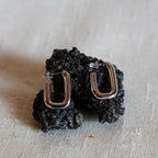 Boucles d'oreilles J'agis, créoles rectangulaires en acier inoxydable argenté, photographiée en vue rapprochée, posées sur une pierre volcanique noire.