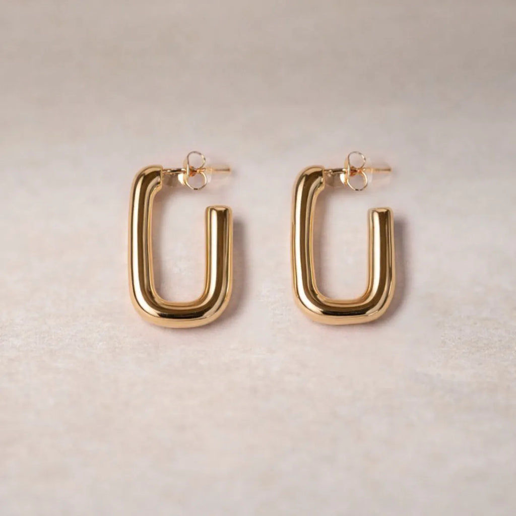 Boucles d'oreilles Franche, créoles rectangulaires en acier inoxydable doré, photographiée en vue rapprochée,  sur une toile beige texturée.