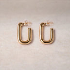 Boucles d'oreilles Franche, créoles rectangulaires en acier inoxydable doré, photographiée en vue rapprochée,  sur une toile beige texturée.