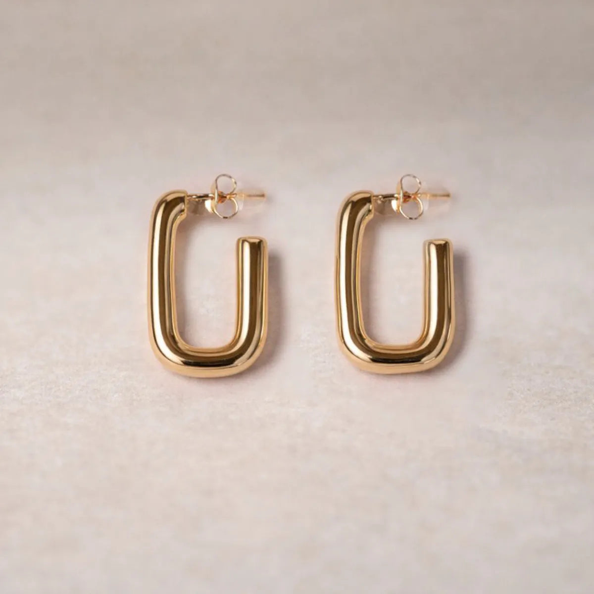 Boucles d'oreilles Franche, créoles rectangulaires en acier inoxydable doré, photographiée en vue rapprochée,  sur une toile beige texturée.