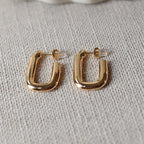 Boucles d'oreilles J'agis, créoles rectangulaires en acier inoxydable doré, photographiée en vue rapprochée, posées sur une toile beige texturée.