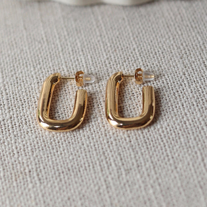 Boucles d'oreilles J'agis, créoles rectangulaires en acier inoxydable doré, photographiée en vue rapprochée, posées sur une toile beige texturée.