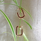 Boucles d'oreilles J'agis, créoles rectangulaires en acier inoxydable doré, photographiée en vue rapprochée, suspendues sur une liane sur une toile beige texturée.