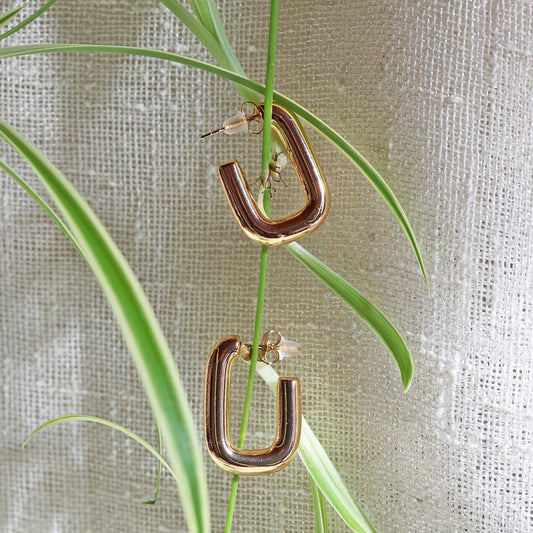 Boucles d'oreilles J'agis, créoles rectangulaires en acier inoxydable doré, photographiée en vue rapprochée, suspendues sur une liane sur une toile beige texturée.