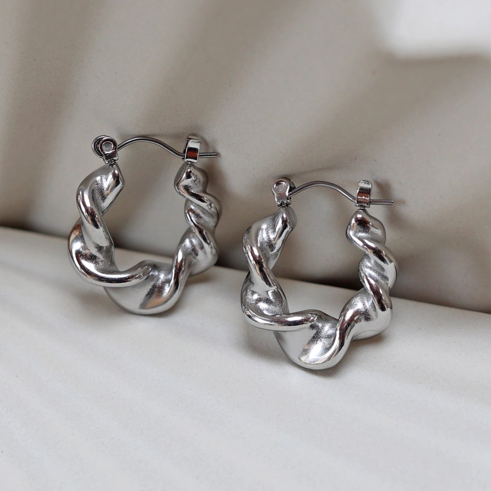 Zoom sur les boucles d’oreilles Elégante - créoles ondulées  argentées en acier inoxydable, présentées sur un support beige texturé.