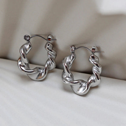 Zoom sur les boucles d’oreilles Elégante - créoles ondulées  argentées en acier inoxydable, présentées sur un support beige texturé.