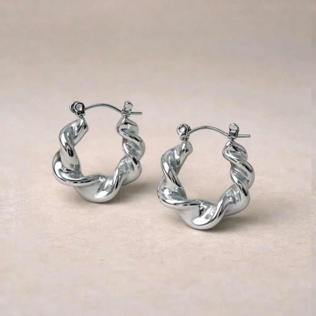 Zoom sur les boucles d’oreilles Elégante - créoles ondulées  argentées en acier inoxydable, présentées sur un fond beige texturé.