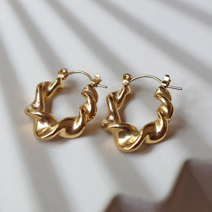 Zoom sur les boucles d’oreilles Elégante - créoles dorées ondulées en acier inoxydable, posées sur un support beige texturé.