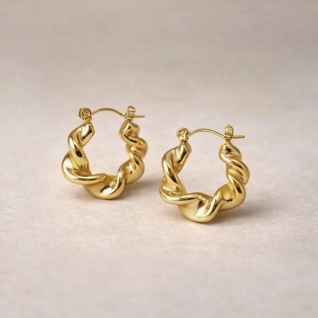 Zoom sur les boucles d’oreilles Elégante - créoles ondulées  dorées en acier inoxydable, présentées sur un fond beige texturé.