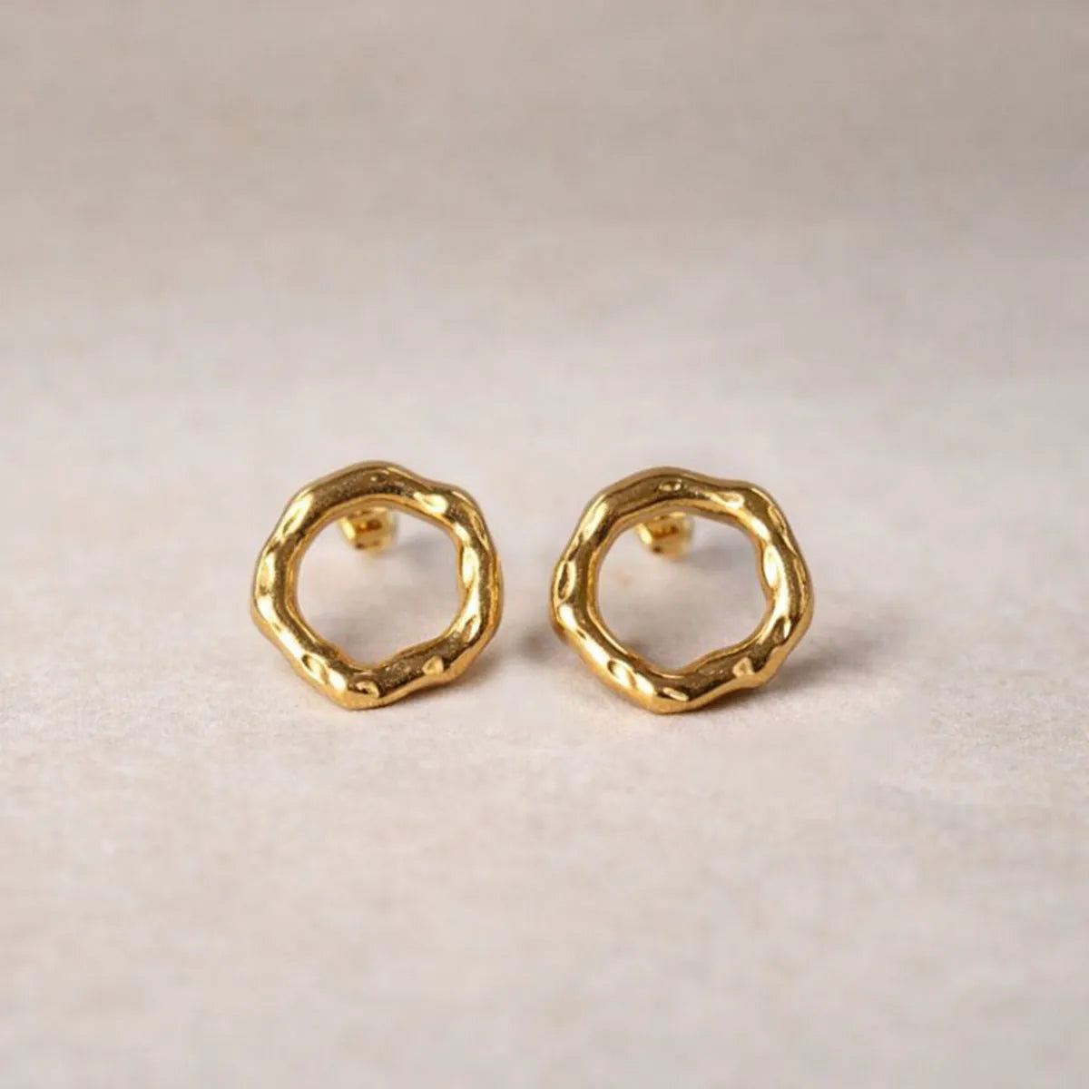 Zoom sur les boucles d'oreilles martelées Naturelle en acier inoxydable doré, posées sur un fond texturé beige.