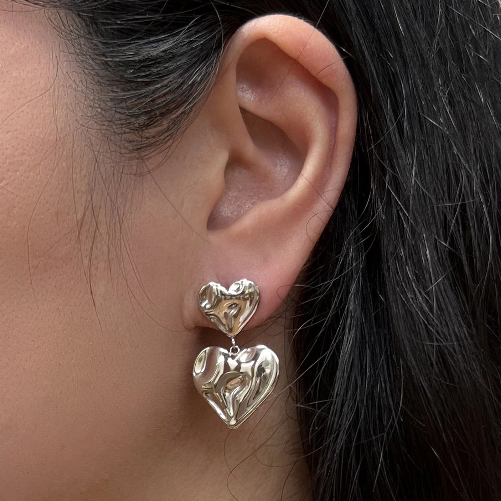Boucles d’oreilles pendantes Grace en acier argenté, ornées de deux coeurs martelés effet ondulé, portées à l'oreille.