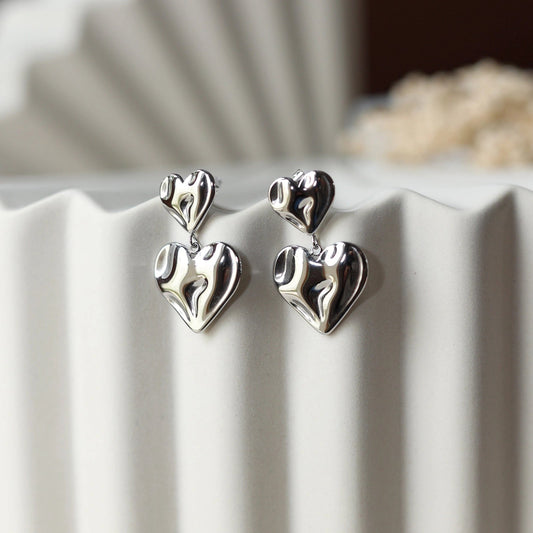 Boucles d’oreilles Grace en acier inoxdydable argenté, design pendantes avec double coeur martelé effet ondulé, posées sur un support texturé beige.