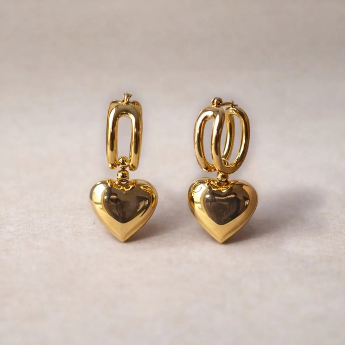 Zoom sur les boucles d'oreilles "Amour suspendu" de Visée, avec pendentifs cœurs en acier doré, sur fond beige texturé.