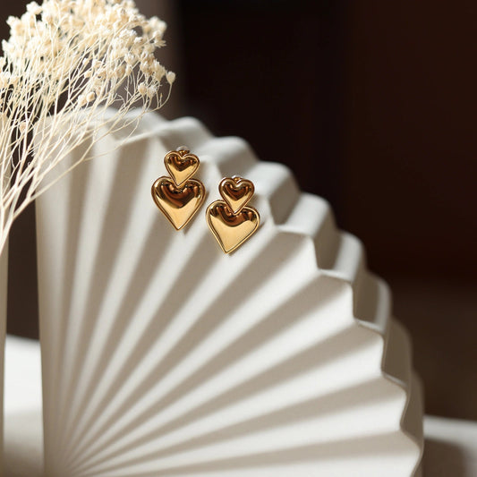 Boucles d'oreilles "Elle m'a dit" en forme de double coeur, en acier doré. Posées sur un fond beige texturé. 