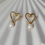 Boucles d’oreilles Loyale, en forme de cœur doré avec perle blanche, en acier inoxydable, photographiées de face sur un fond beige texturé.
