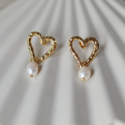 Boucles d’oreilles Loyale, en forme de cœur doré avec perle blanche, en acier inoxydable, photographiées de face sur un fond beige texturé.