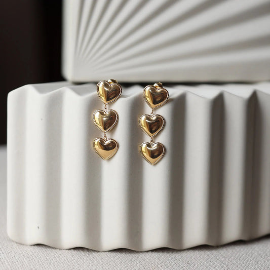 Boucles d'oreilles "Unique", en acier inoxydable doré, pendantes en coeurs, photographiées sur un fond beige texturé.