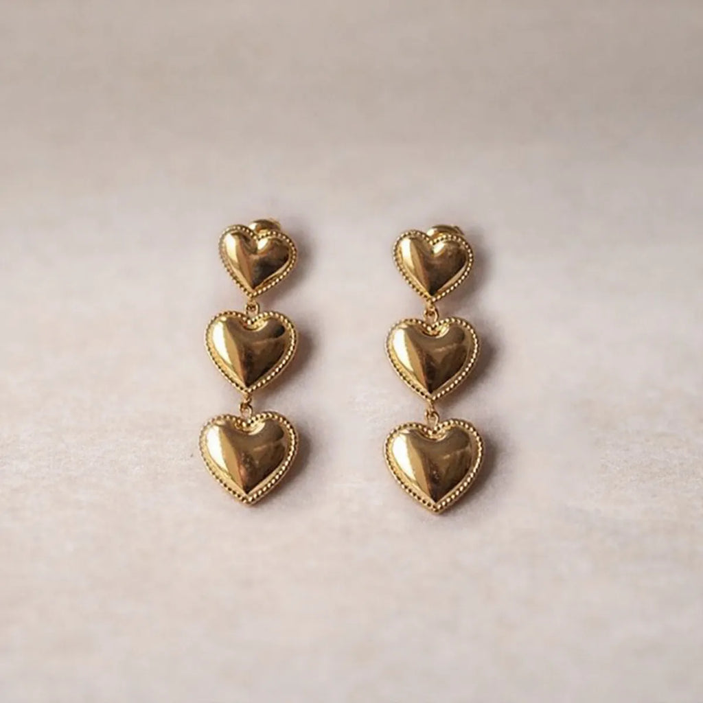 Boucles d'oreilles "Unique", en acier inoxydable doré, pendantes en cœurs, photographiées sur un fond beige texturé.