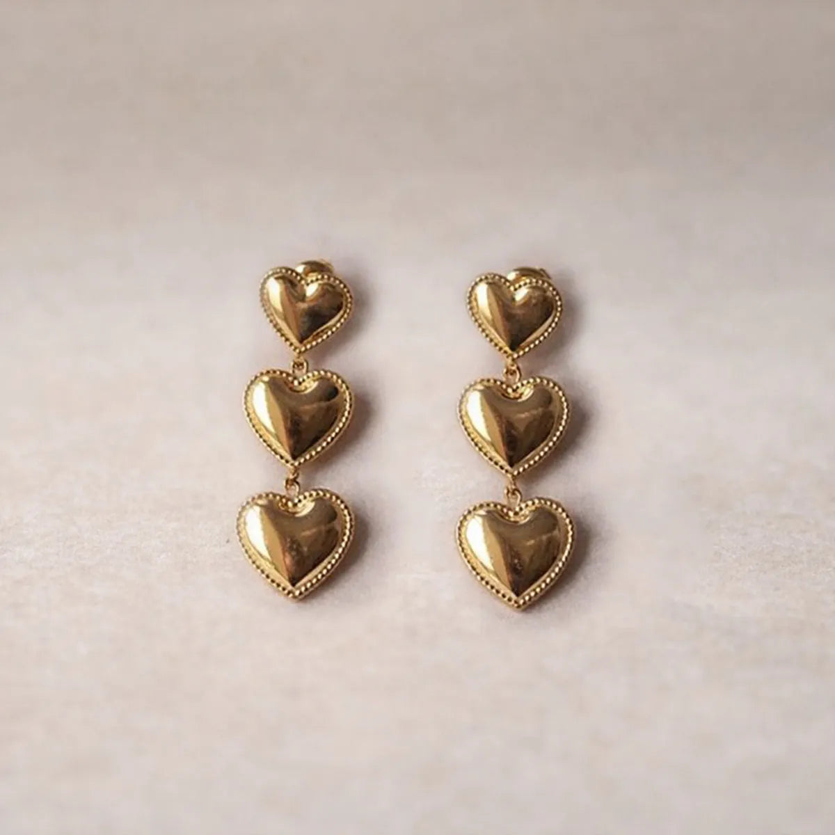 Boucles d'oreilles "Unique", en acier inoxydable doré, pendantes en cœurs, photographiées sur un fond beige texturé.