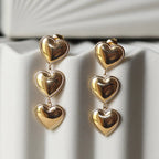 Boucles d'oreilles "Unique", en acier inoxydable doré, pendantes en coeurs, photographiées sur un fond beige texturé.
