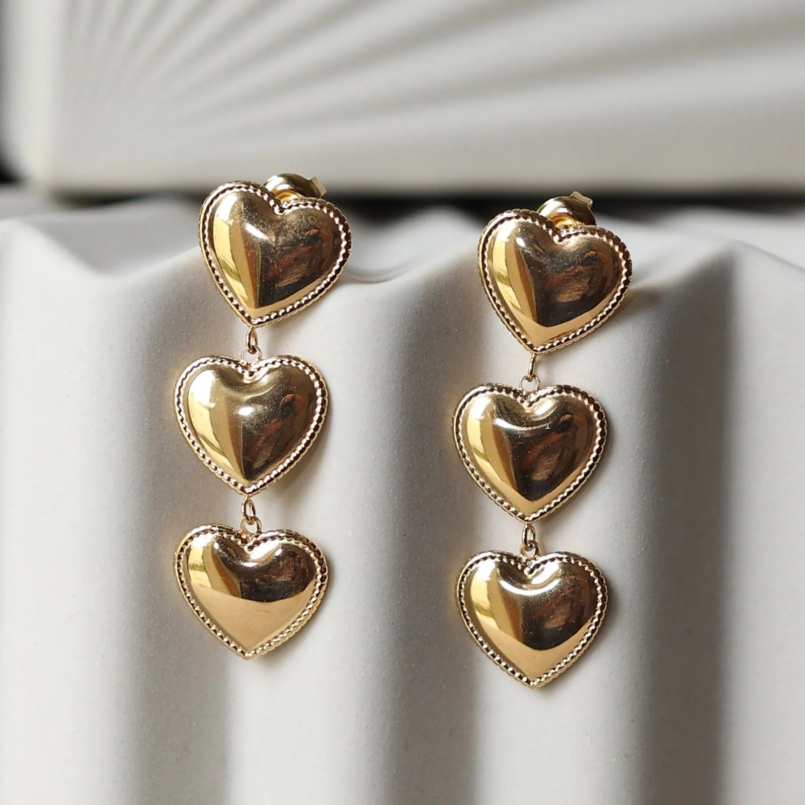 Boucles d'oreilles "Unique", en acier inoxydable doré, pendantes en coeurs, photographiées sur un fond beige texturé.
