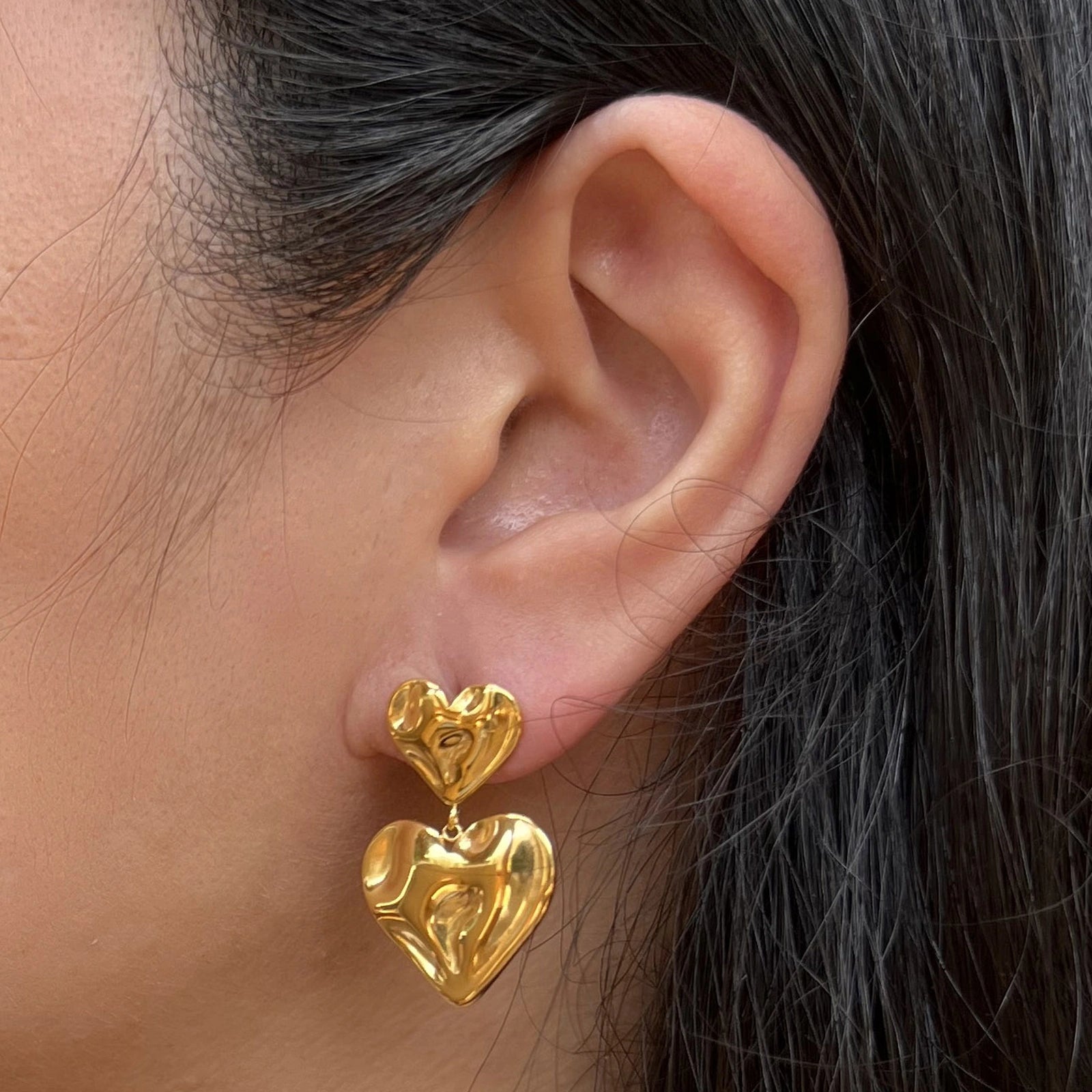 Boucles d’oreilles pendantes Grace en acier doré, ornées de deux coeurs martelés effet ondulé, portées à l'oreille.