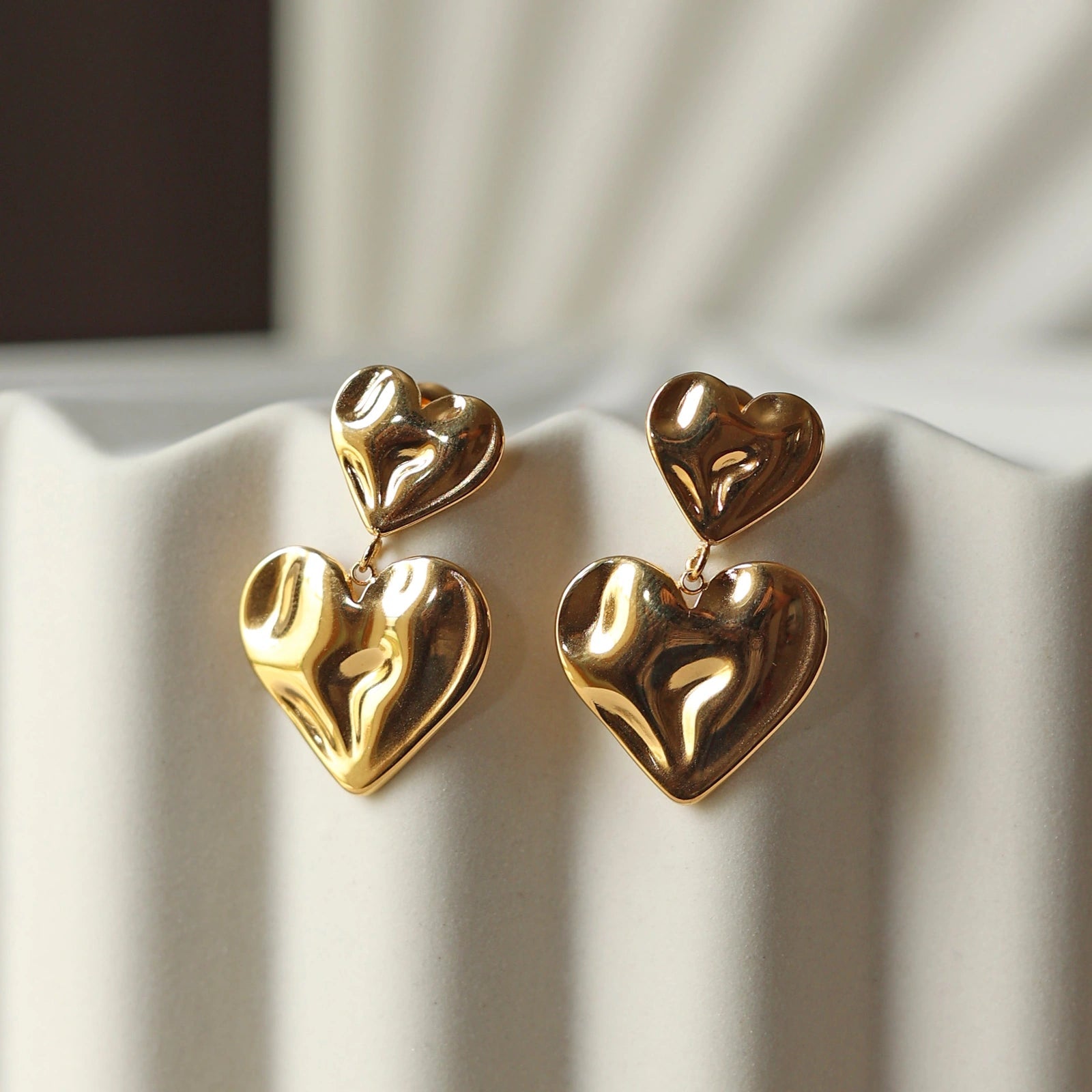 Boucles d’oreilles Grace en acier inoxdydable doré, design pendantes avec double coeur martelé effet ondulé, posées sur un support texturé beige.