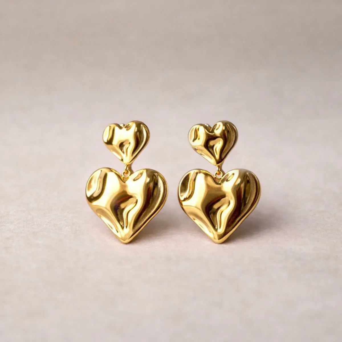 Boucles d’oreilles Grace en acier inoxdydable doré, design pendantes avec double coeur martelé effet ondulé, posées sur fond texturé beige.