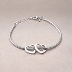 Zoom sur le bracelet "Coeurs" en acier argenté, orné de deux cœurs mobiles, posé sur un fond beige texturé.