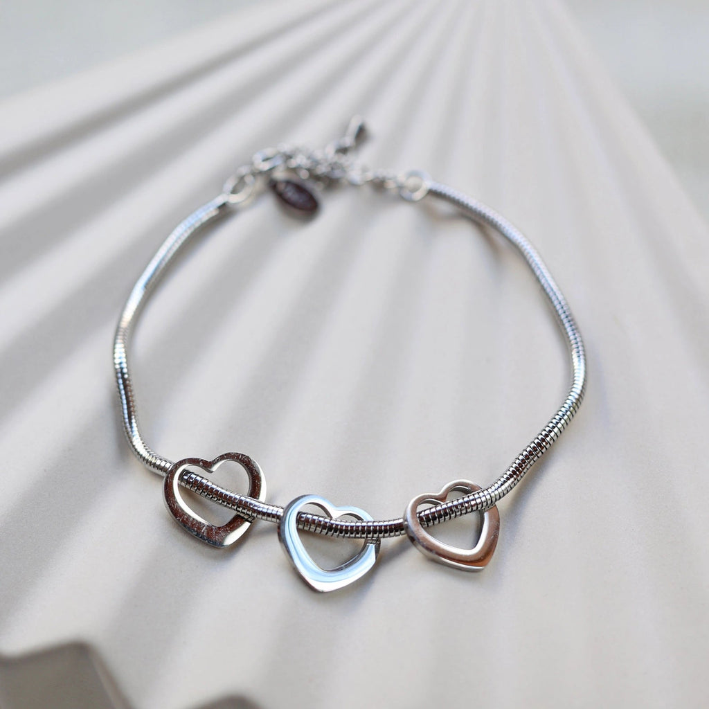 Zoom sur le bracelet "Emotive" en acier argenté, orné de trois cœurs mobiles, posé sur un fond beige texturé.
