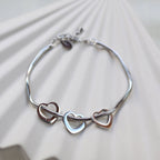 Zoom sur le bracelet "Emotive" en acier argenté, orné de trois cœurs mobiles, posé sur un fond beige texturé.