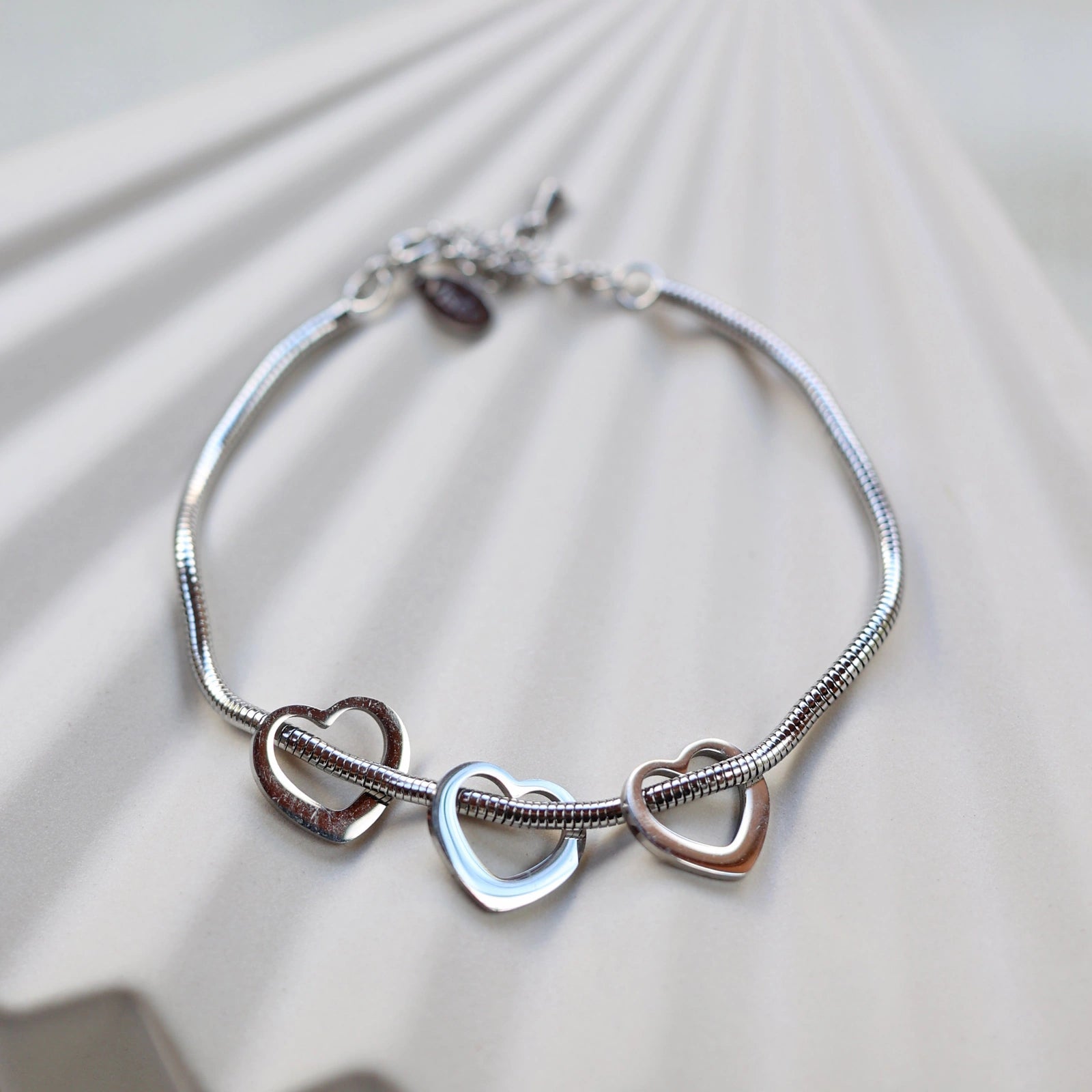 Zoom sur le bracelet "Emotive" en acier argenté, orné de trois cœurs mobiles, posé sur un fond beige texturé.
