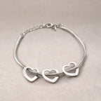 Zoom sur le bracelet "Coeurs" en acier argenté, orné de trois cœurs mobiles, posé sur un fond beige texturé.