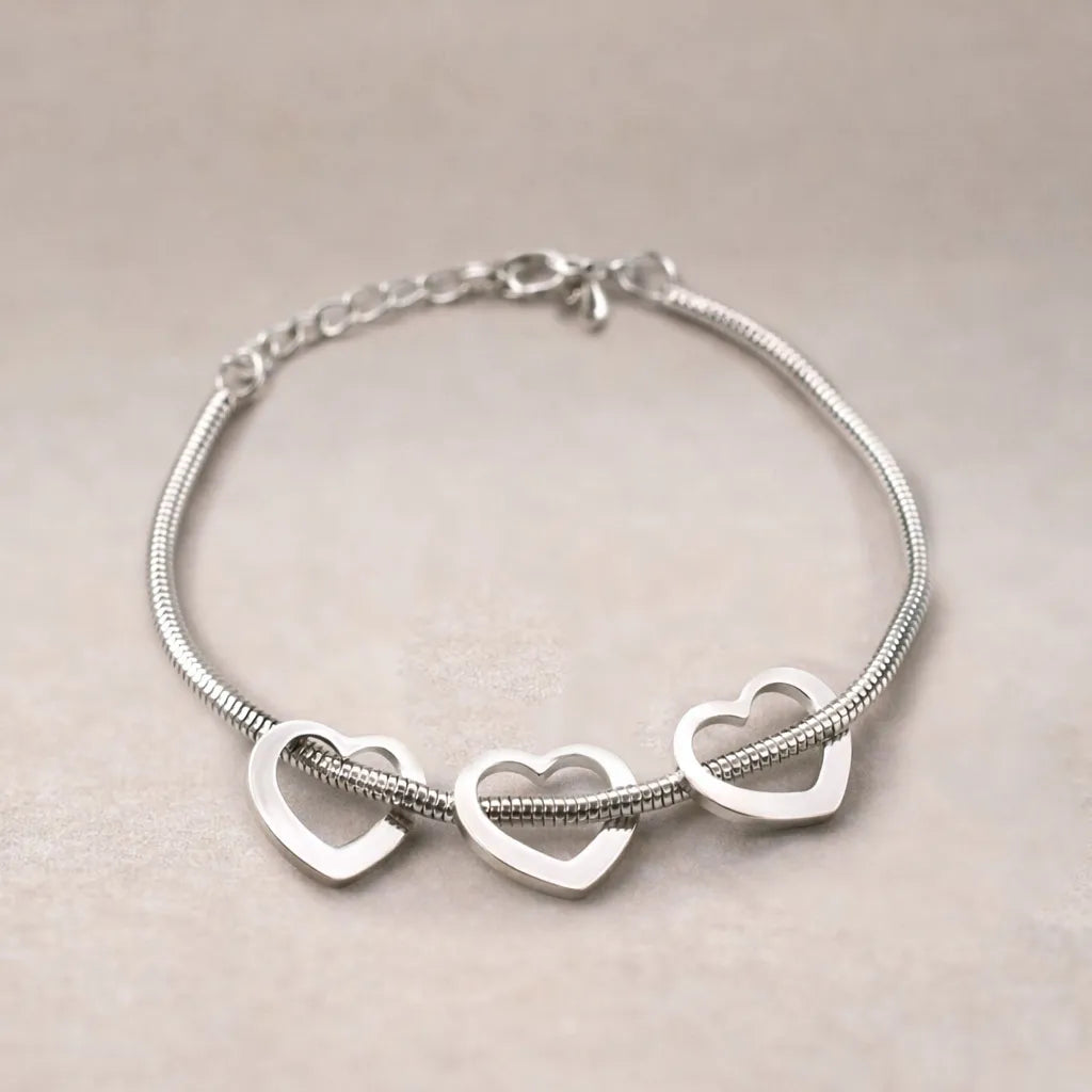 Zoom sur le bracelet "Coeurs" en acier argenté, orné de trois cœurs mobiles, posé sur un fond beige texturé.