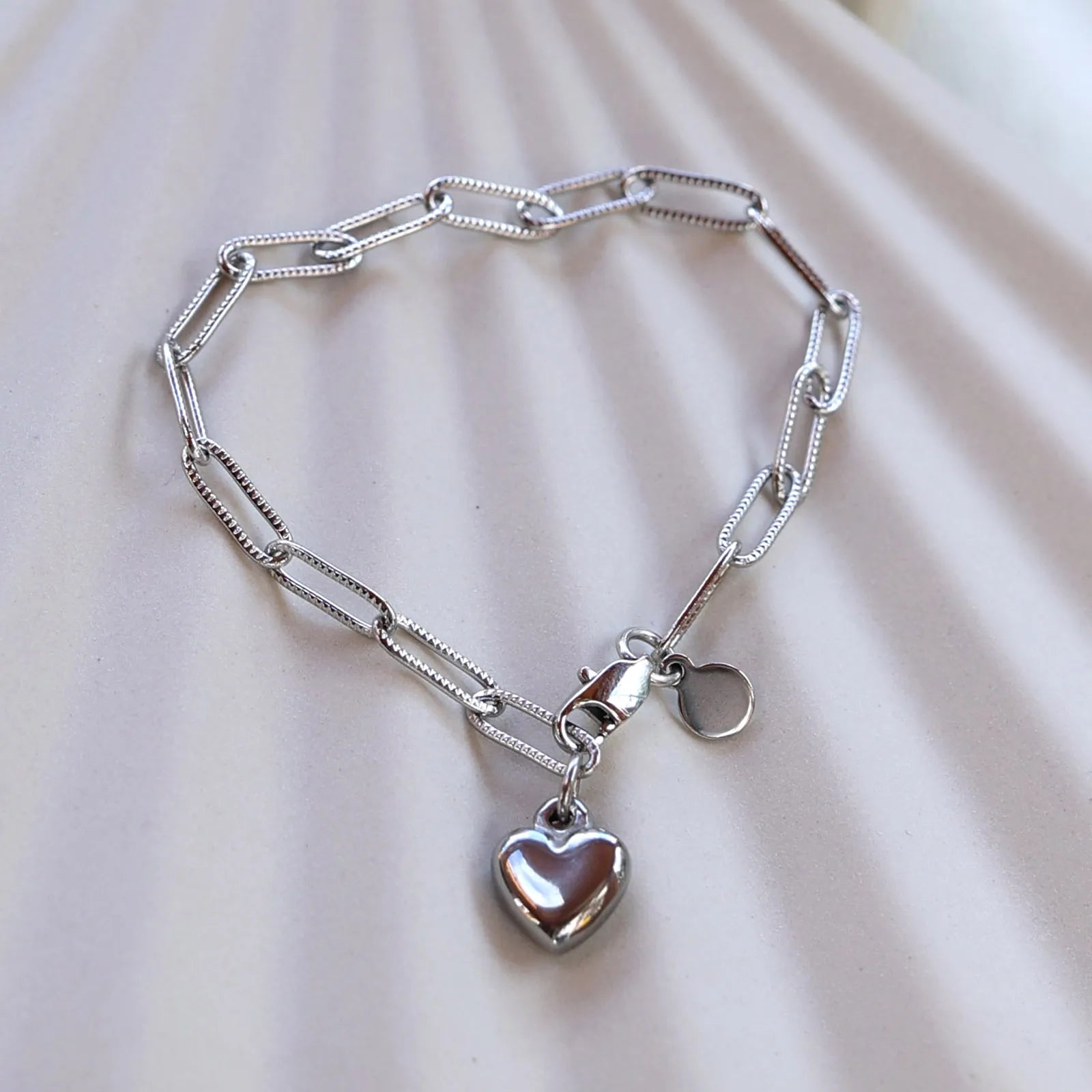 Bracelet Valeur acier inoxydable argenté, avec breloque cœur personnalisable, posé sur un fond beige texturé.