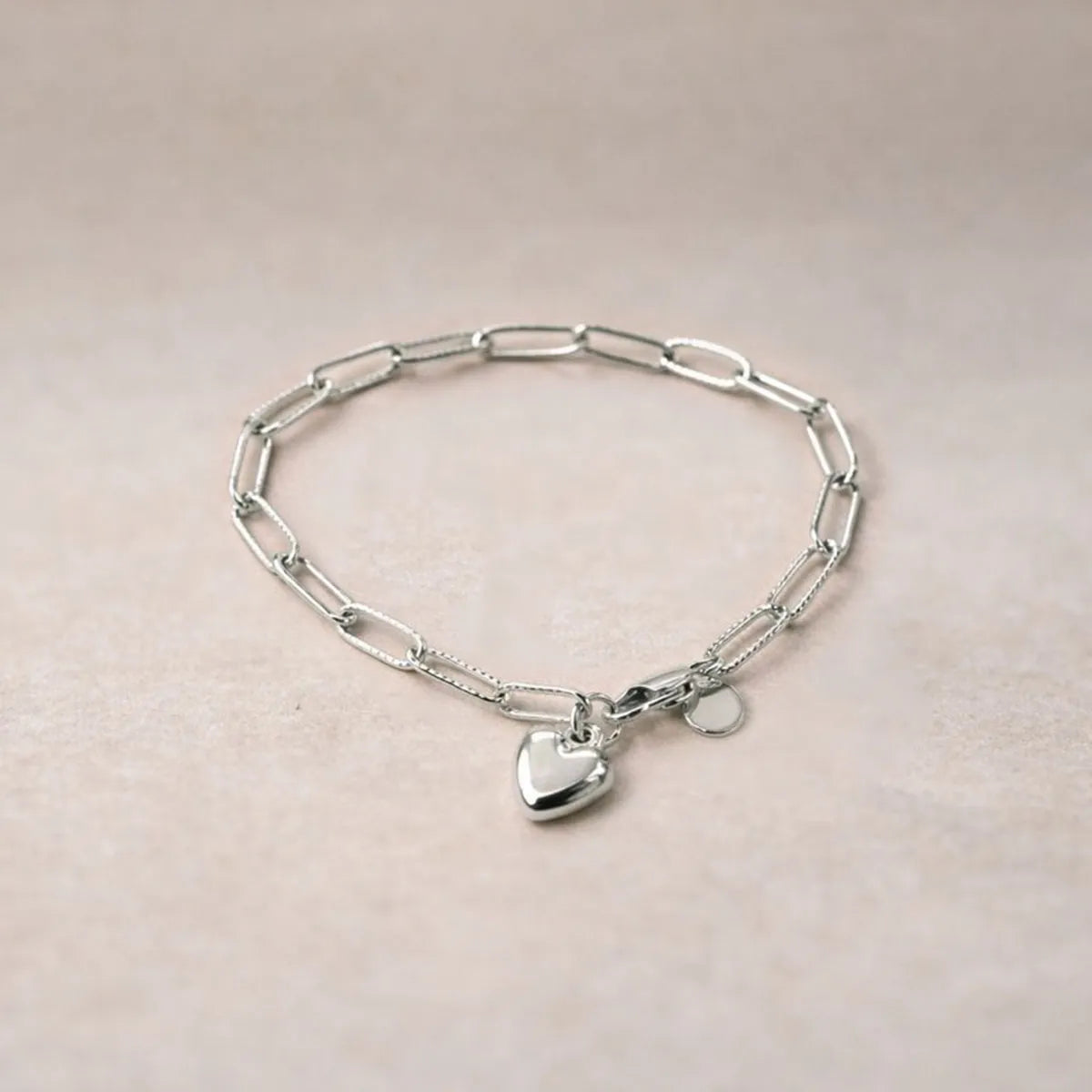 Bracelet Valeur acier inoxydable argenté, avec breloque cœur personnalisable, posé sur un fond beige texturé.