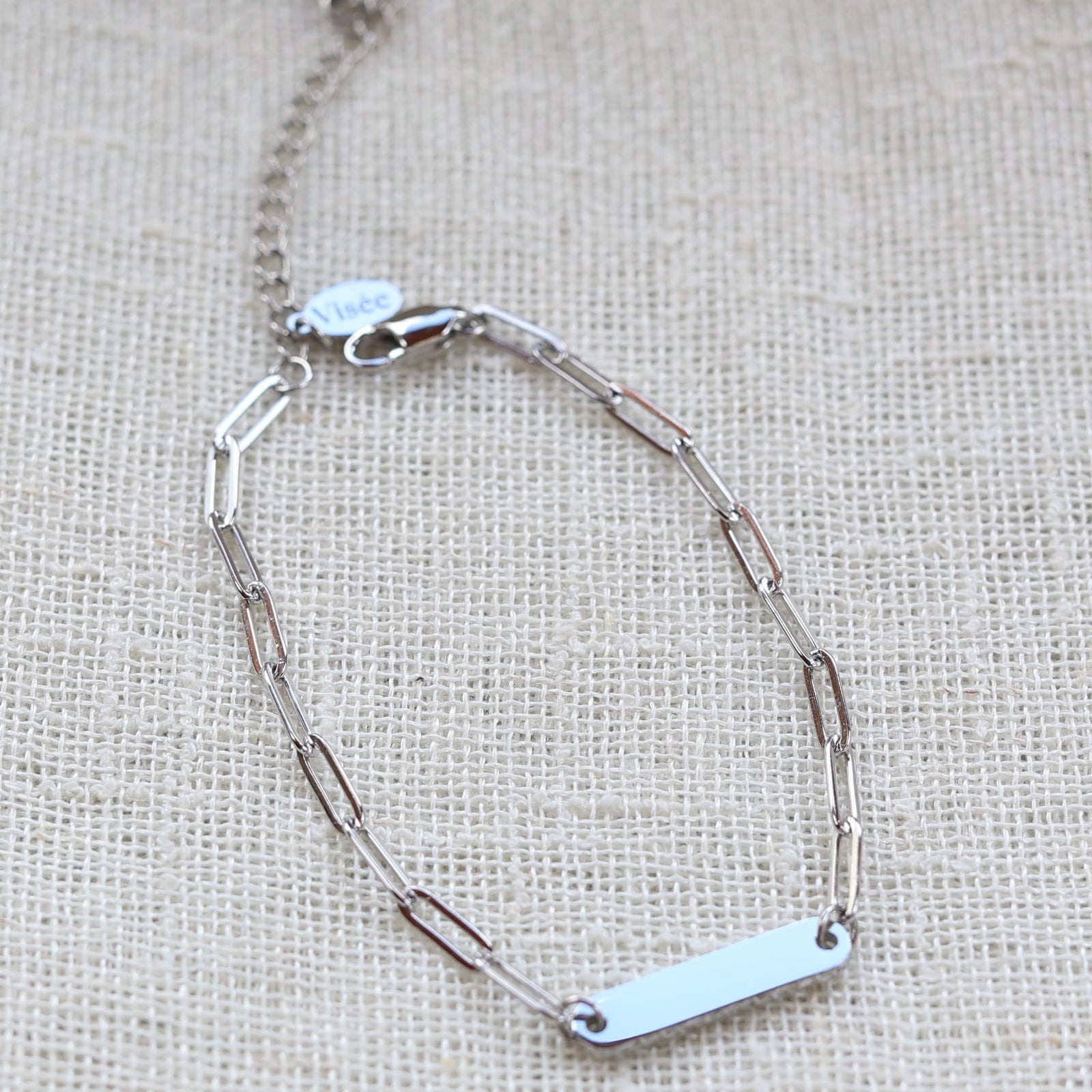 Bracelet "Expression" en acier inoxydable argenté, à personnaliser avec une plaque gravable, présenté sur un fond beige texturé.