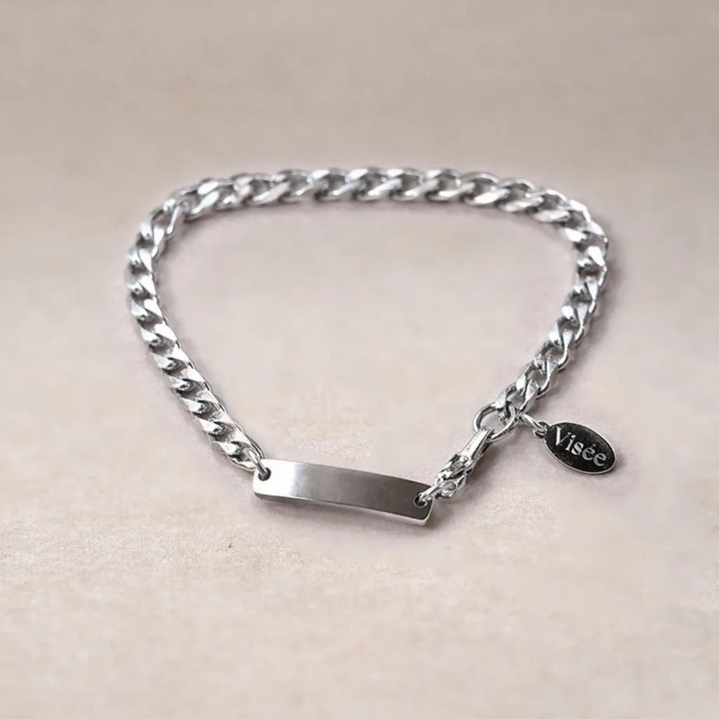 Bracelet Force en acier argenté, style gourmette, avec plaque gravable, posé sur un fond beige texturé et petite breloque Visée