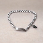 Bracelet Force en acier argenté, style gourmette, avec plaque gravable, posé sur un fond beige texturé et petite breloque Visée