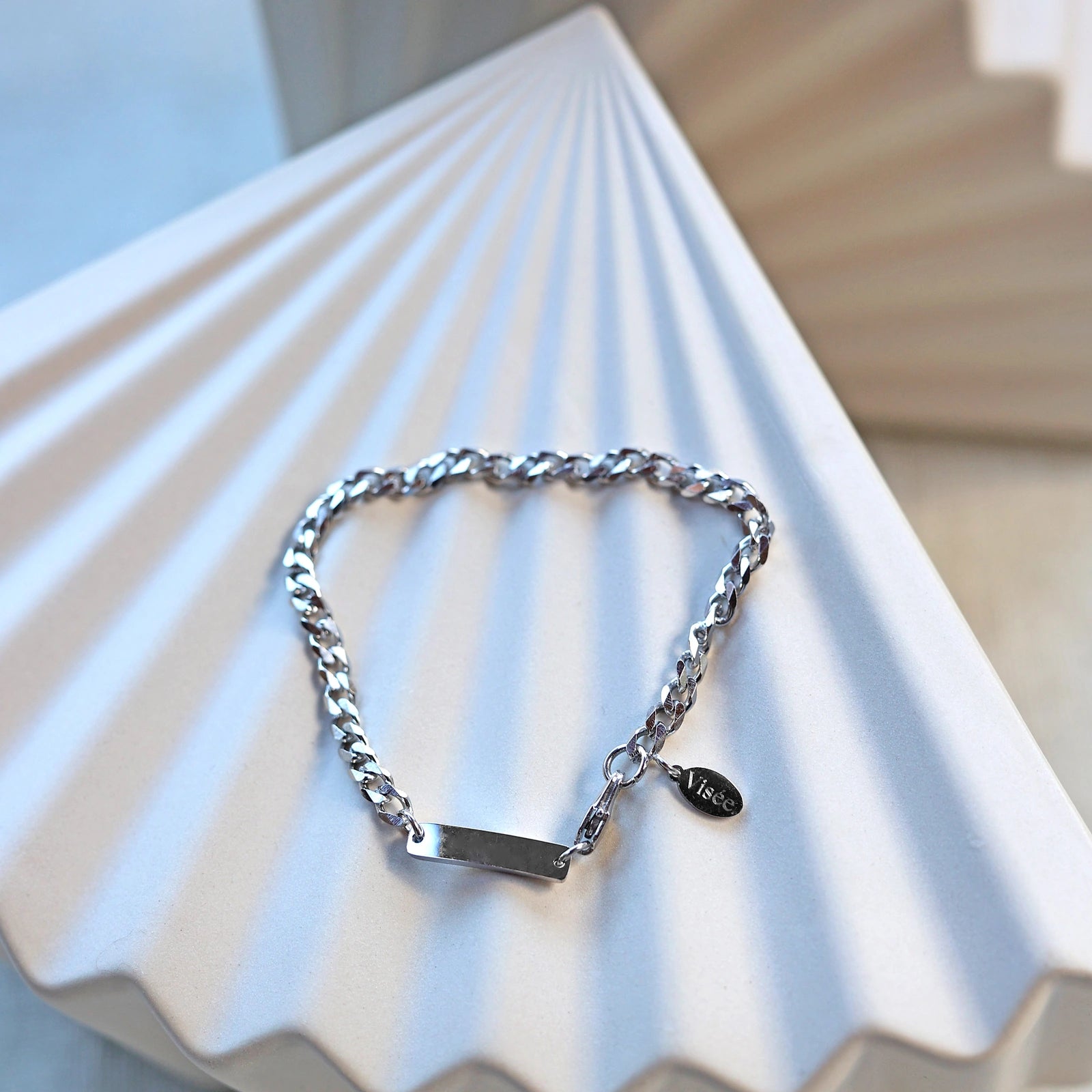 Bracelet "Libère toi" en acier argenté, style gourmette, avec plaque gravable, posé sur un fond beige texturé.