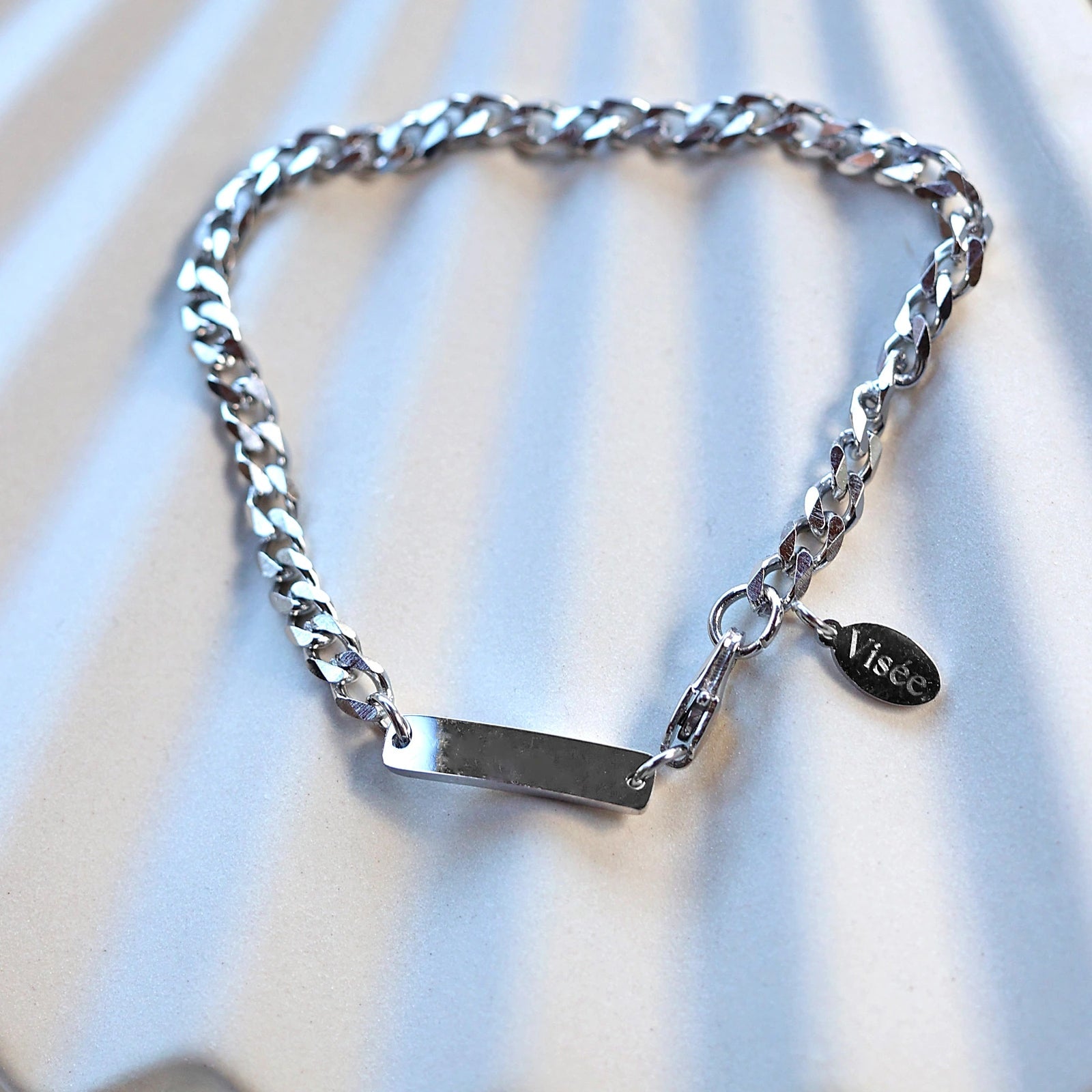 Zoom sur le bracelet "Libère toi" en acier argenté, style gourmette, avec plaque gravable, posé sur un fond beige texturé.