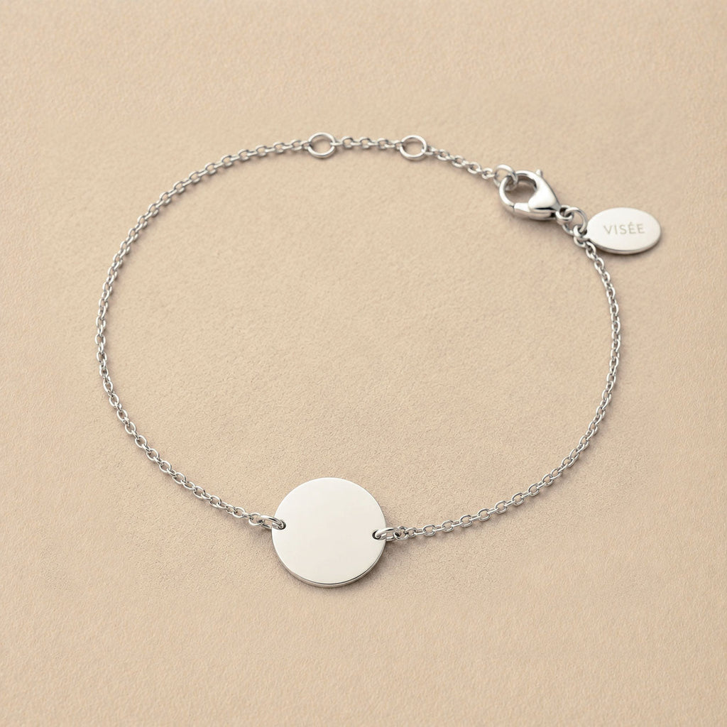 Zoom sur le bracelet "Aimé" en acier argenté personnalisable sur les 2 faces avec médaille ronde et une chaine fine , présenté sur un tissu beige texturé. 
