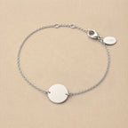 Zoom sur le bracelet "Aimé" en acier argenté personnalisable sur les 2 faces avec médaille ronde et une chaine fine , présenté sur un tissu beige texturé. 