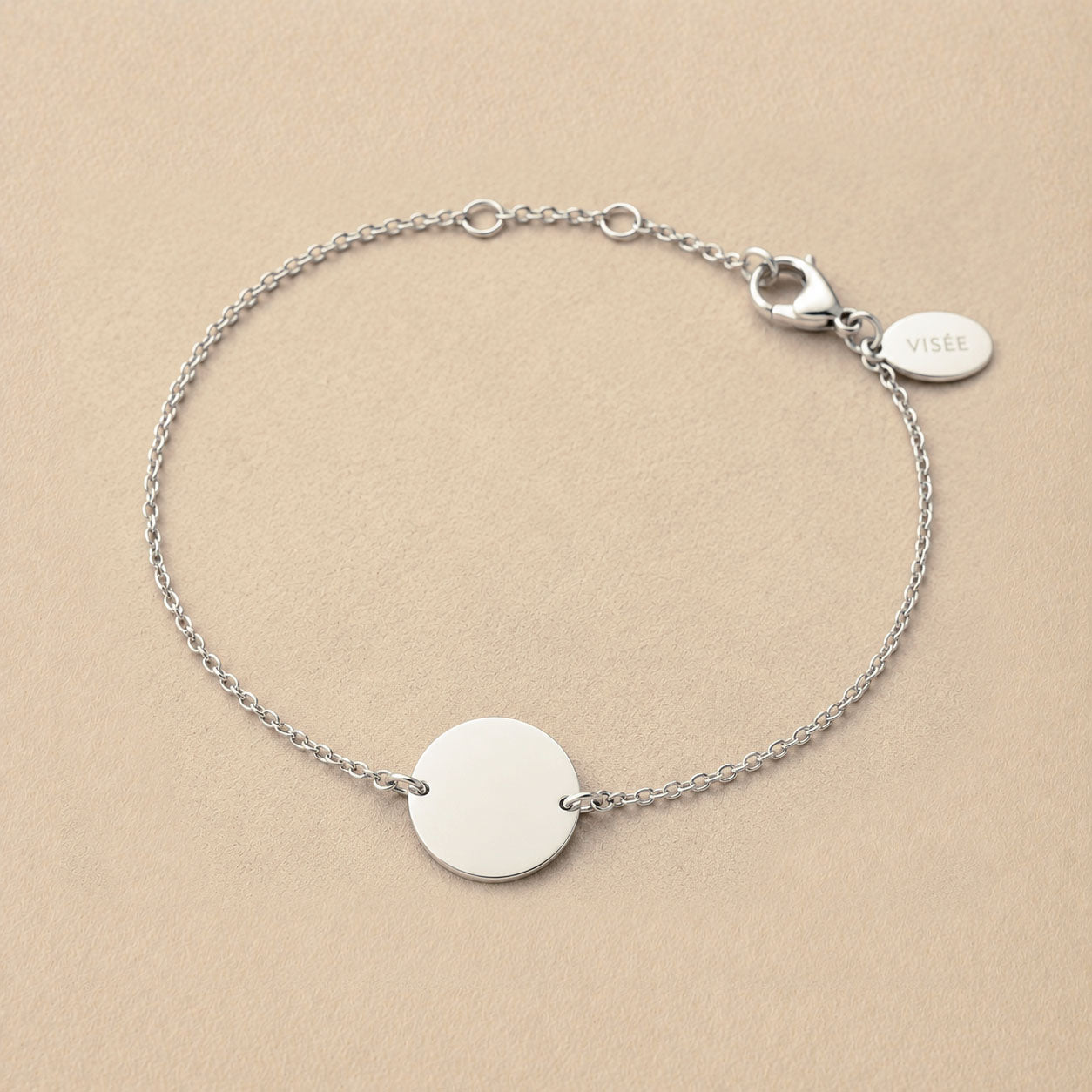 Zoom sur le bracelet "Aimé" en acier argenté personnalisable sur les 2 faces avec médaille ronde et une chaine fine , présenté sur un tissu beige texturé. 