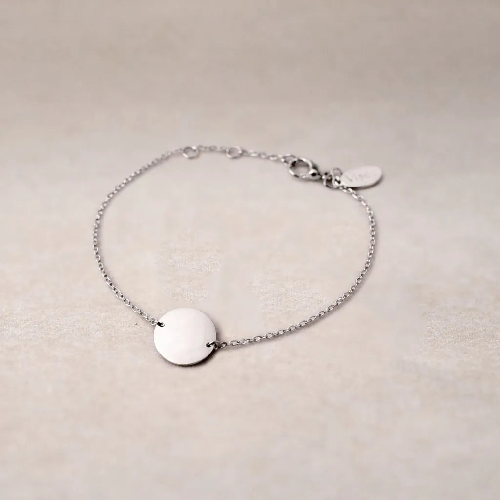 Zoom sur le bracelet "Confiance" en acier argenté personnalisable avec médaille ronde et une chaine fine , présenté sur un tissu beige texturé. 